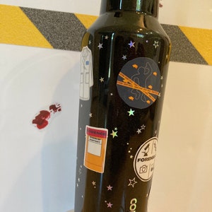 May include: Black stainless steel water bottle with a black glitter finish and gold stars. The bottle has several stickers on it, including a lab coat, a crime scene tape sticker, a sticker that says "Evidence, Skin of Suspect", and a sticker that says "Forensics". The bottle is a Corkcicle brand.