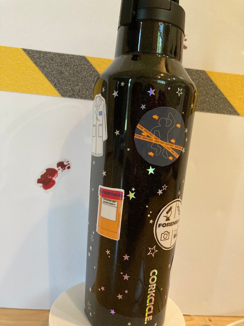 May include: Black stainless steel water bottle with a black glitter finish and gold stars. The bottle has several stickers on it, including a lab coat, a crime scene tape sticker, a sticker that says "Evidence, Skin of Suspect", and a sticker that says "Forensics". The bottle is a Corkcicle brand.