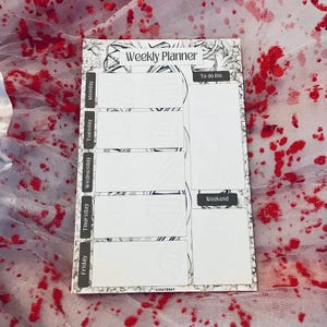 May include: A white weekly planner with a black floral border and the words "Weekly Planner" at the top. The planner has sections for each day of the week, a to-do list, and a weekend section. The text "LICIA TODAY" is at the bottom.