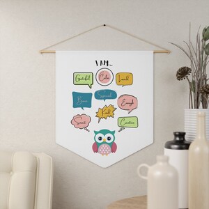 Può includere: Un banner bianco con le parole "I AM..." e affermazioni positive in fumetti colorati. Un'illustrazione di un gufo è in basso. Il banner è sospeso da un'asta di legno con una corda.