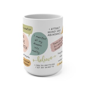 May include: White ceramic mug with colorful text bubbles containing affirmations like "I manifest all my dreams into reality" and "I attract money and abundance." The word "believe" is written in green script.