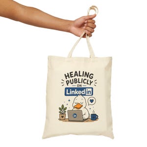 Puede incluir: Bolso tote color crema con el texto "HEALING PUBLICLY on LinkedIn". Presenta un pato de dibujos animados trabajando en un portátil con el logotipo de LinkedIn, una planta, una taza de café y un icono de corazón.