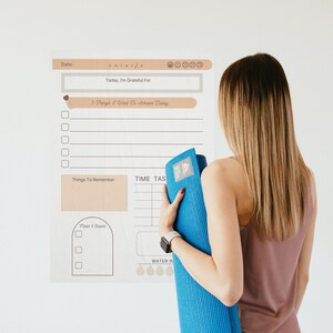 May include: A white printable planner with a brown header and a section for daily gratitude, things to remember, time tasks, and water intake. The planner is on a white wall with a woman holding a blue yoga mat.