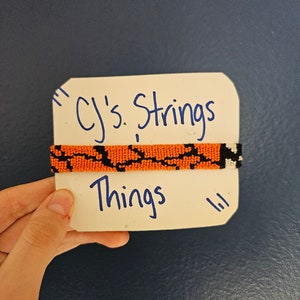 May include: An orange and black beaded bracelet is displayed on a white card. The card has the text "CJ's Strings Things" written in blue ink. The bracelet is held by a hand against a dark blue background.