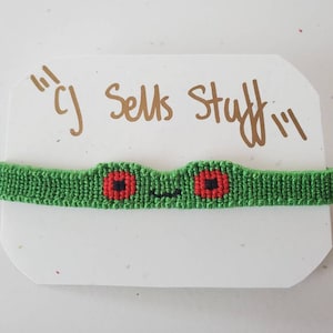 May include: A green macrame bracelet featuring a frog face design with red and black details. The bracelet is displayed on a white card with the text "cj Sells Stuff" in gold lettering.