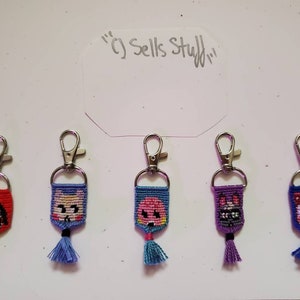 May include: Five handmade keychains with cartoon character designs. Each keychain features a silver clasp, a woven design with a cartoon face, and a tassel. The keychains are red, blue, pink, purple, and blue.