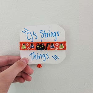 May include: Handmade orange friendship bracelet with a Halloween theme. The bracelet features a repeating pattern of black cats, purple bats, and green and white accents. The text "CJ's Strings Things" is written on a white card.