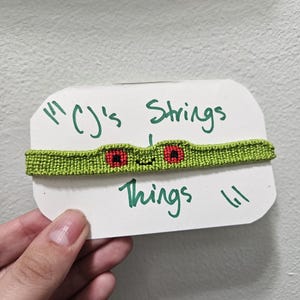 May include: A green woven friendship bracelet featuring a frog face design with two red eyes and a small black smile. The bracelet is attached to a white rectangular display card with the handwritten text CJ's Strings and Things in green ink.