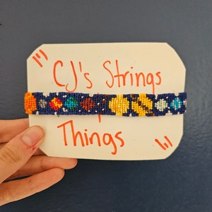 May include: A handmade friendship bracelet with a dark blue background and colorful geometric patterns. The bracelet features orange, yellow, brown, and blue designs. The text "CJ's Strings Things" is written on a white card.