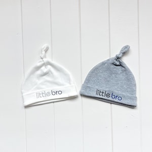 May include: Two baby hats, one white and one gray, with the text "little bro" embroidered on each.