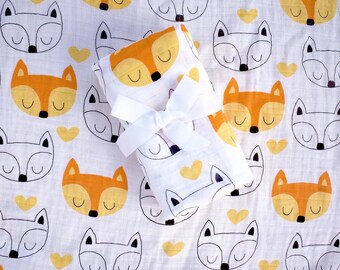 May include: A white baby blanket with a repeating pattern of orange and white foxes and yellow hearts. The blanket is folded and tied with a white ribbon. The blanket is made of a soft, lightweight material.