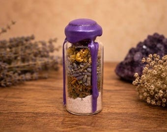 May include: A clear glass bottle filled with layers of dried chamomile flowers, lavender buds, green herbs, and white salt. The bottle is sealed with a purple wax cap that has drips running down the sides of the glass container.