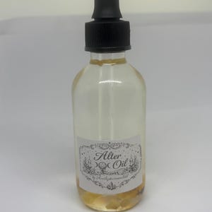 May include: A clear glass bottle with a black dropper top, filled with a light yellow liquid. The bottle has a white label with the words "Alter Oil" and decorative floral elements. The bottom of the bottle contains small, yellow stones.