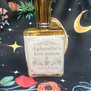 May include: A perfume bottle with a gold cap and a white label that reads "Aphrodite's love potion." The label has decorative floral and heart designs. The bottle is rectangular with a gold rim and is filled with a light amber liquid.