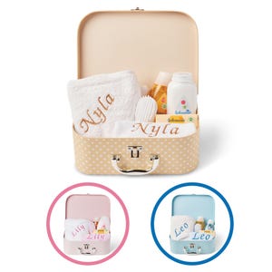 May include: A beige suitcase with a white polka dot pattern and a silver handle. The suitcase is open and contains a white towel with the name "Nyla" embroidered on it, a white washcloth with the name "Nyla" embroidered on it, a bottle of baby lotion, a bottle of baby powder, a baby brush, and a bar of soap.