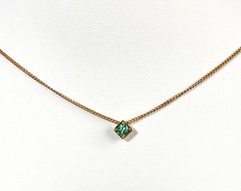 May include: A delicate gold-toned necklace with a small, square, emerald-green gemstone pendant. The necklace is displayed on a white mannequin, showcasing the minimalist design and the vibrant color of the gemstone.