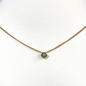 Puede incluir: Un delicado collar dorado con un pequeño colgante cuadrado de piedra preciosa verde esmeralda. El collar se exhibe en un maniquí blanco, mostrando el diseño minimalista y el color vibrante de la piedra preciosa.
