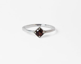 May include: A silver ring featuring a square, dark red gemstone. The gemstone is set in a square bezel, and the ring band is a simple, polished silver. The ring is displayed on a white background.
