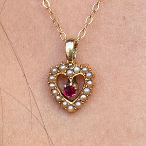 May include: A gold chain necklace with a heart-shaped pendant. The pendant is made of gold and has a small red gemstone in the centre. The pendant is surrounded by small white pearls.