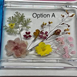 Peut inclure: Pièce d'art en résine rectangulaire transparente avec des fleurs séchées. Les fleurs sont dans des tons de rose, jaune et marron, avec le texte "Option A" en noir.