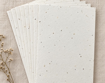 May include: A stack of rectangular off-white paper sheets with a textured surface containing small, scattered dark and light flecks. The paper is arranged in a fan-like formation, showcasing the fibrous, handmade appearance of each individual sheet.