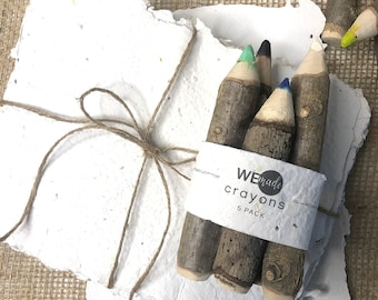 May include: A stack of white handmade paper with deckled edges tied with brown twine, accompanied by a five-pack of twig-shaped crayons wrapped in a paper band labelled WE made crayons 5 PACK, with additional loose twig crayons nearby.