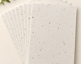 May include: A stack of rectangular sheets of off-white handmade paper with a textured surface and visible dark flecks throughout. The paper is arranged in a fan shape, showcasing the deckled edges of each individual sheet in the collection.