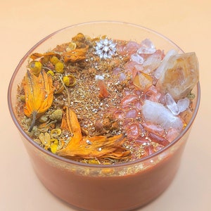 Op de afbeelding: Een bruine kaars met gedroogde bloemen, kristallen en glitter ingebed in de was. De kaars zit in een glazen pot.