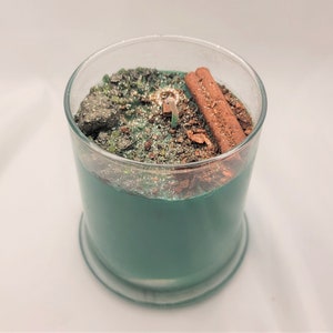 May include: A teal green candle with a glass jar and a silver lid. The candle has a layer of cinnamon sticks and glitter on top.