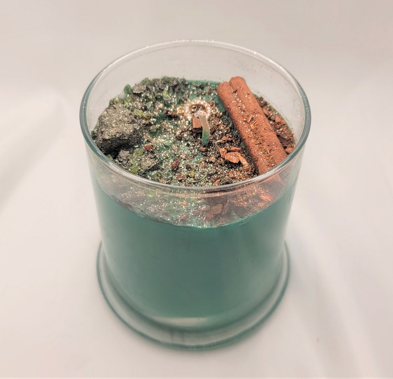 May include: A teal green candle with a glass jar and a silver lid. The candle has a layer of cinnamon sticks and glitter on top.