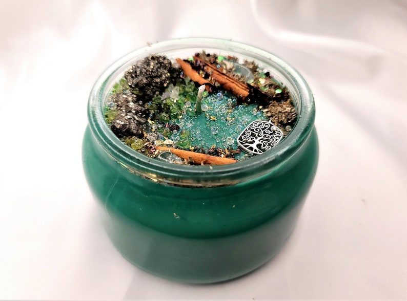 May include: A green glass jar candle with a silver tree of life charm, cinnamon sticks, and green and gold glitter.