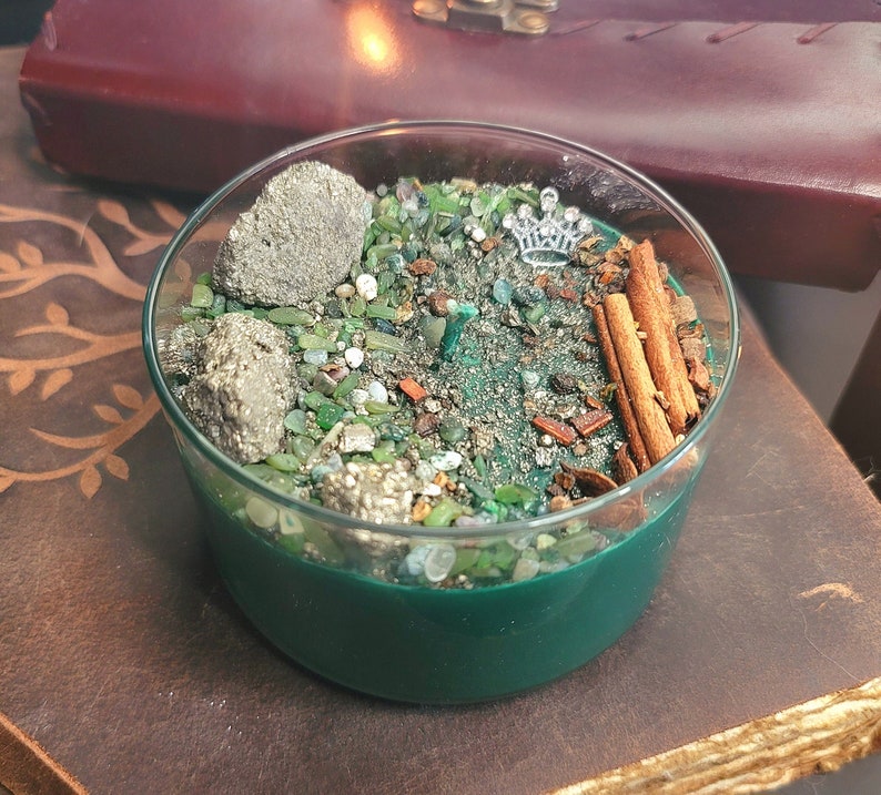 May include: A green glass candle with a crown design on top. The candle is filled with green and gold glitter, small rocks, and cinnamon sticks.