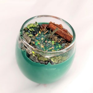May include: A teal green candle in a glass jar with a variety of natural elements embedded in the wax, including cinnamon sticks, green and brown crystals, and glitter.