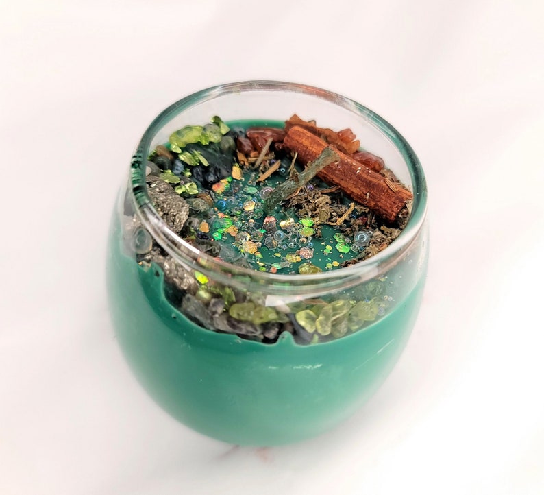 May include: A teal green candle in a glass jar with a variety of natural elements embedded in the wax, including cinnamon sticks, green and brown crystals, and glitter.