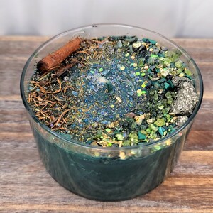 May include: A teal green candle with a clear glass jar. The candle is topped with a variety of natural elements, including cinnamon sticks, dried herbs, and small green and gold stones.