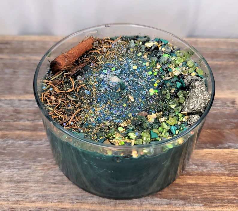 May include: A teal green candle with a clear glass jar. The candle is topped with a variety of natural elements, including cinnamon sticks, dried herbs, and small green and gold stones.