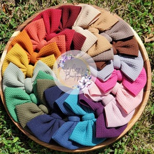 May include: A wooden tray filled with a variety of colorful fabric bows. The bows are arranged in a circular pattern, showcasing a spectrum of colors including red, orange, yellow, green, blue, and purple. The bows have a textured appearance.