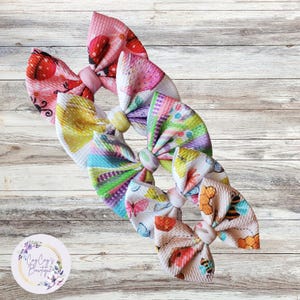 May include: Four colorful hair bows with different floral and insect patterns. The bows are made of a soft, textured fabric and are tied together with a white ribbon.