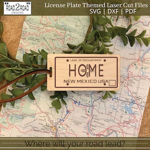 May include: A wooden sign with a brown background and white text that reads "LAND OF ENCHANTMENT" and "HOME" with a stylized graphic of the state of New Mexico. The text "NEW MEXICO USA" is also on the sign. The sign is on top of a map of the state of New Mexico.