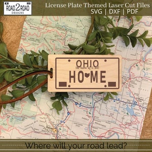 Könnte beinhalten: Eine Holz-Lizenzplatten-Themen-Laserdatei mit dem Text "OHIO the heart of it all! HOME". Die Platte ist an einer braunen Lederschnur befestigt. Der Hintergrund umfasst eine Karte und Grünzeug.