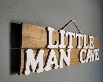 Puede incluir: Letrero de madera con las palabras "LITTLE MAN CAVE" en letras blancas en relieve. El letrero está hecho de dos tablas de madera con un acabado marrón claro, y está suspendido por una cuerda natural. El letrero está montado en una pared gris.