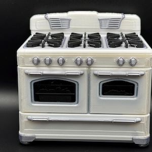 May include: A miniature white stove with a silver trim and black burner grates. The appliance features six silver control knobs on the front panel, two oven doors with black windows, and a raised back panel with two silver vent-like details.