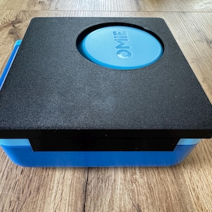 May include: A blue and black lunchbox with a round, light blue lid. The lid has the word "OMIE" embossed on it. The lunchbox is rectangular with a black top and a blue base. The lunchbox is sitting on a wooden surface.
