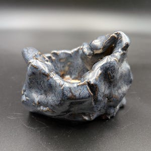 May include: A small, hand-formed ceramic bowl with an irregular, craggy shape. The exterior features a mottled blue and grey glaze with dark, metallic-toned accents along the ridges and crevices, creating a textured, rock-like appearance.