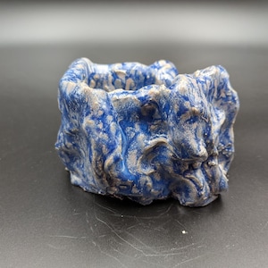 May include: A small, hand-formed ceramic vessel with an irregular, organic shape. The surface features a marbled, swirled pattern of deep royal blue and metallic silver glaze, creating a textured, uneven appearance with raised ridges and deep crevices.