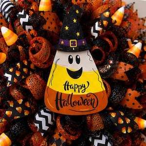 May include: A Halloween wreath with a candy corn centerpiece wearing a witch hat. The candy corn has a smiling face and the words "Happy Halloween". The wreath is decorated with orange, black, and white ribbons and candy corn accents.
