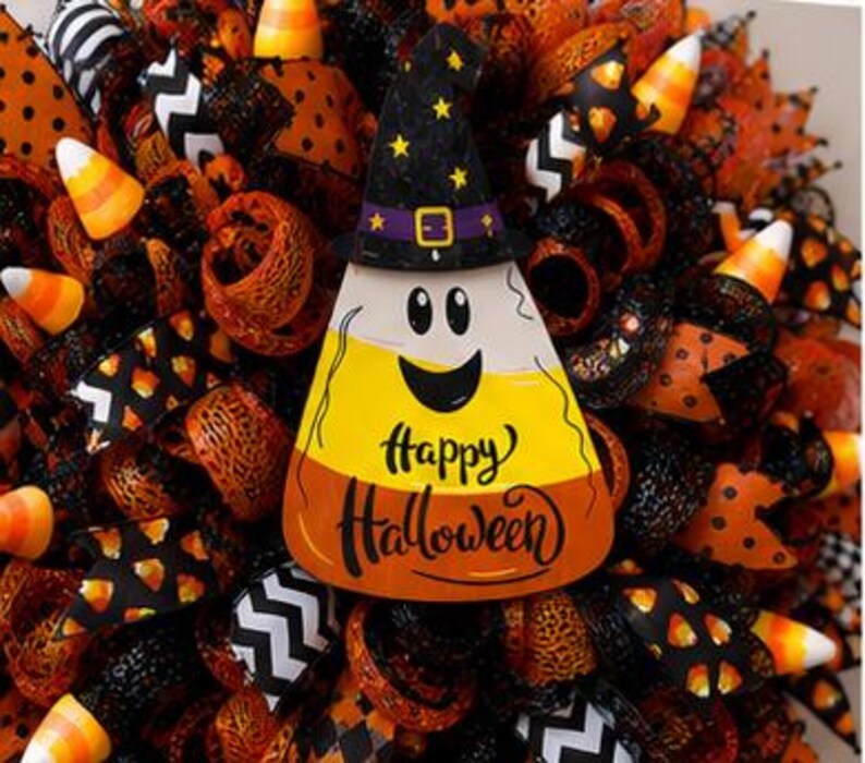 May include: A Halloween wreath with a candy corn centerpiece wearing a witch hat. The candy corn has a smiling face and the words "Happy Halloween". The wreath is decorated with orange, black, and white ribbons and candy corn accents.