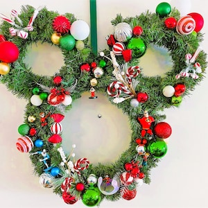 Puede incluir: Una corona de Navidad con forma de cabeza de Mickey Mouse, decorada con adornos rojos, verdes y blancos, bastones de caramelo y pequeñas figuras.