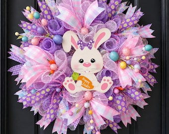 May include: A decorative wreath featuring purple and pink mesh, plaid ribbons, and small colourful speckled eggs. A central cutout of a white bunny with a purple polka dot bow holds an orange carrot with the text Happy Easter printed on it.