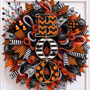 Peut inclure: Une couronne décorative d'Halloween composée de rubans en maille noirs, orange et blancs avec des motifs à rayures, à carreaux et à pois. Le centre affiche le mot BOO en lettres en bois superposées avec des motifs chevron, rayés et à pois.